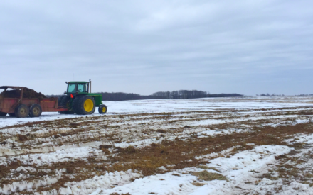 A tractor pulls a wagon spreading manure across a field covered with snow