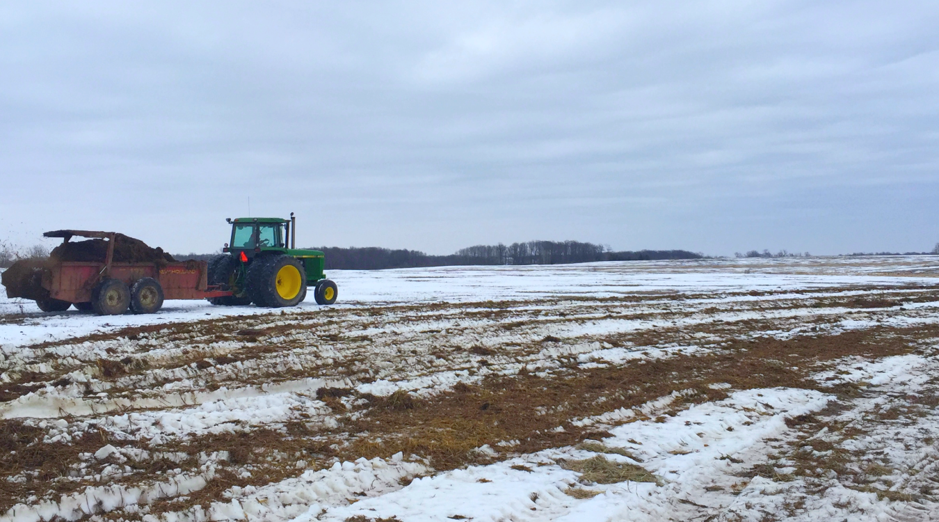 A tractor pulls a wagon spreading manure across a field covered with snow