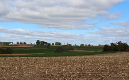 A rural American agricultural landscape. In the foreground is a harvested field with corn stubble remaining. Beyond that are rolling hills with patches of green farmland and some trees scattered throughout. In the distance, a small farm community is visible with several buildings including farm structures with silos.