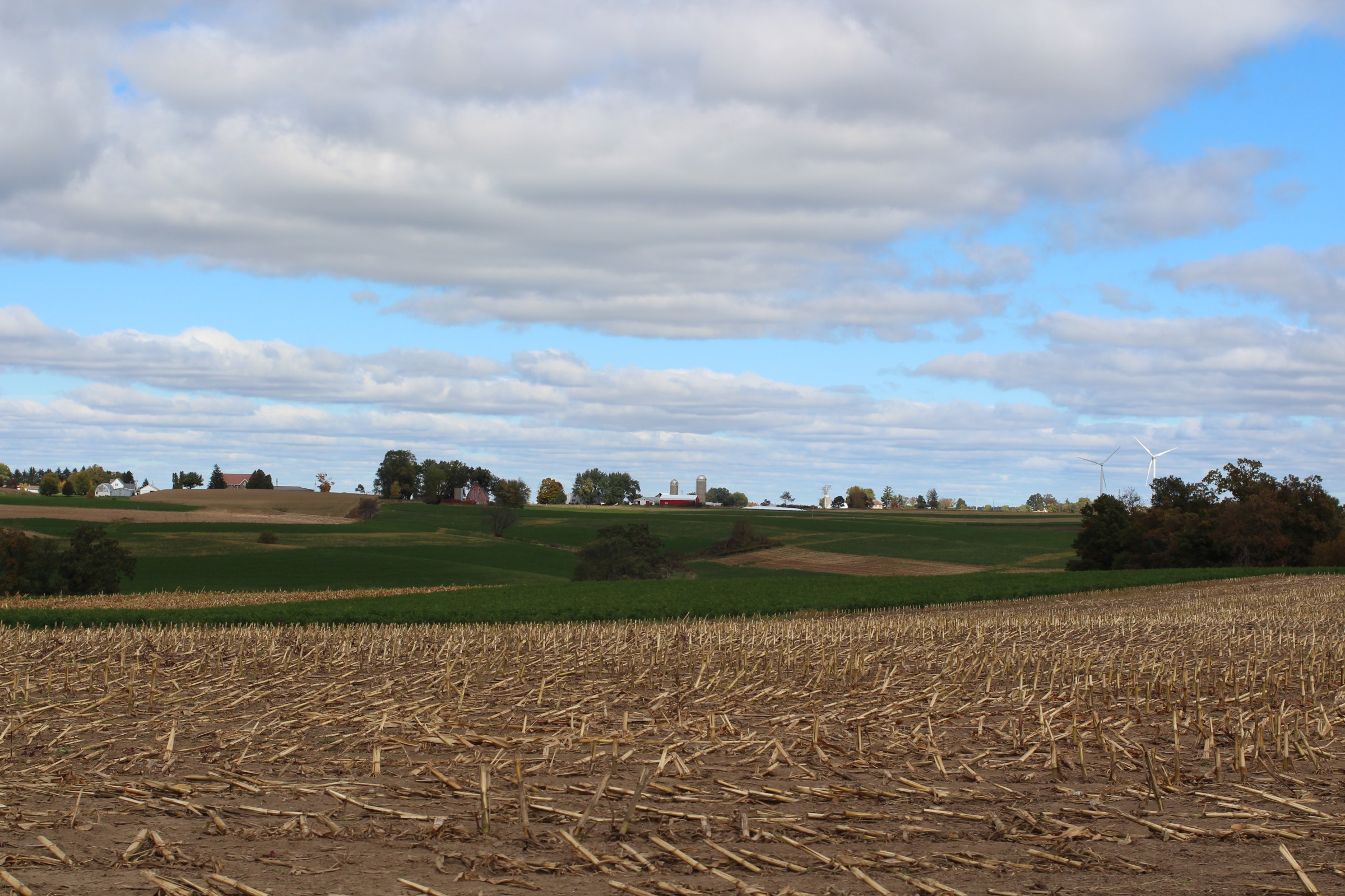 A rural American agricultural landscape. In the foreground is a harvested field with corn stubble remaining. Beyond that are rolling hills with patches of green farmland and some trees scattered throughout. In the distance, a small farm community is visible with several buildings including farm structures with silos.