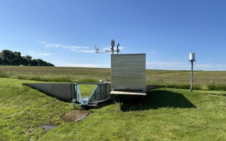 Water-monitoring station with metal flume, sensors, and a small weather instrument tower in a grassy field under a clear sky.