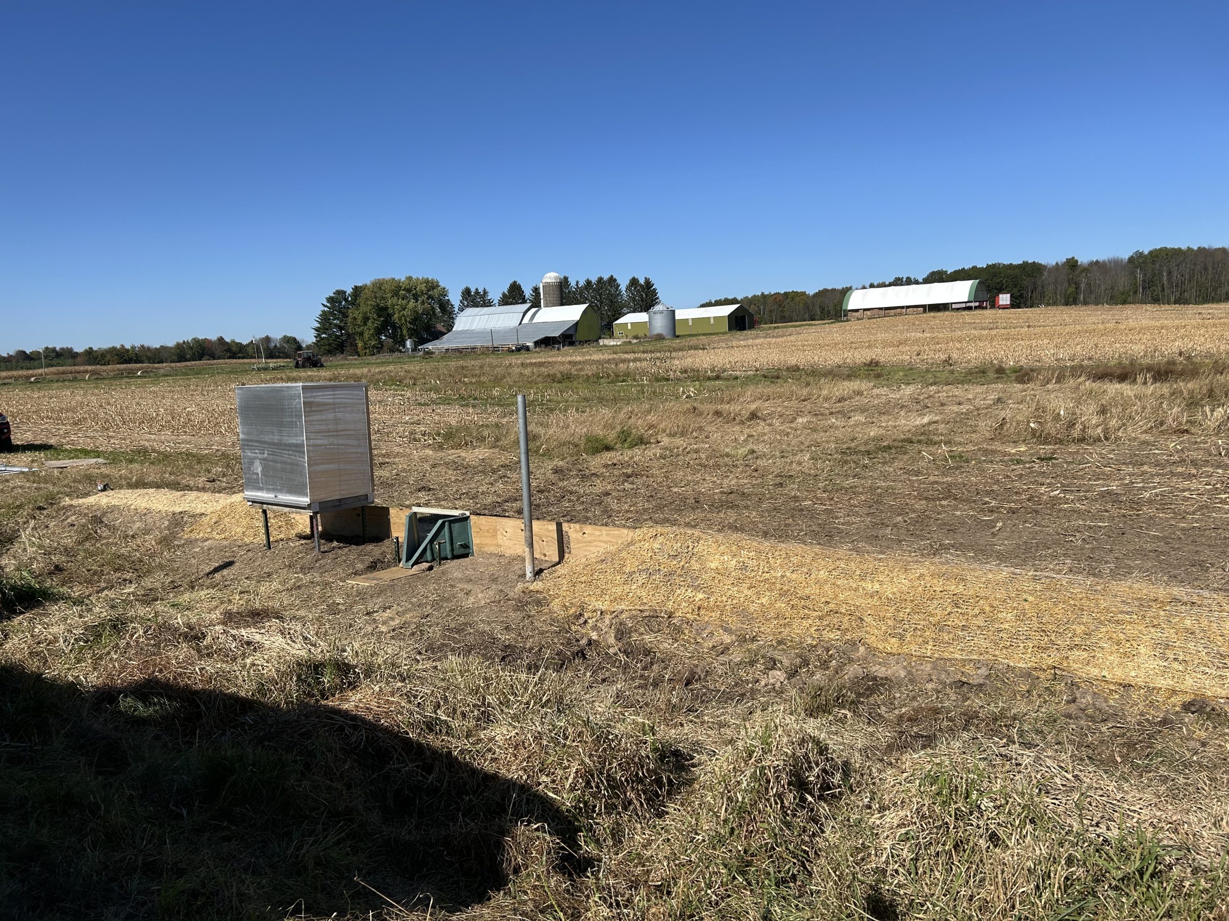 Runoff monitoring station with flume in harvested field with farm buildings in background